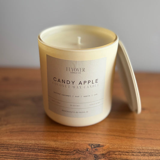 A creamy off-white coconut wax candle in a matching glass jar with a lid leaning against it, placed on a wooden surface against a neutral gray background. The label reads 'FLOVER CANDLE CO and fragrance is Candy Apple.