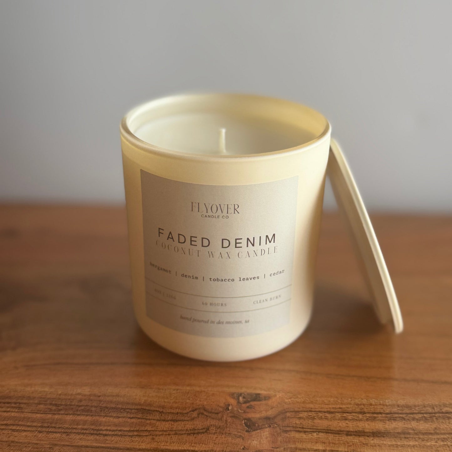 A creamy off white coconut wax candle in an ivory vessel with the lid leaning on the side. A fragrance label of Faded Denim and Flyover Candle Co. Candle is on top of a wooden table with white back drop.