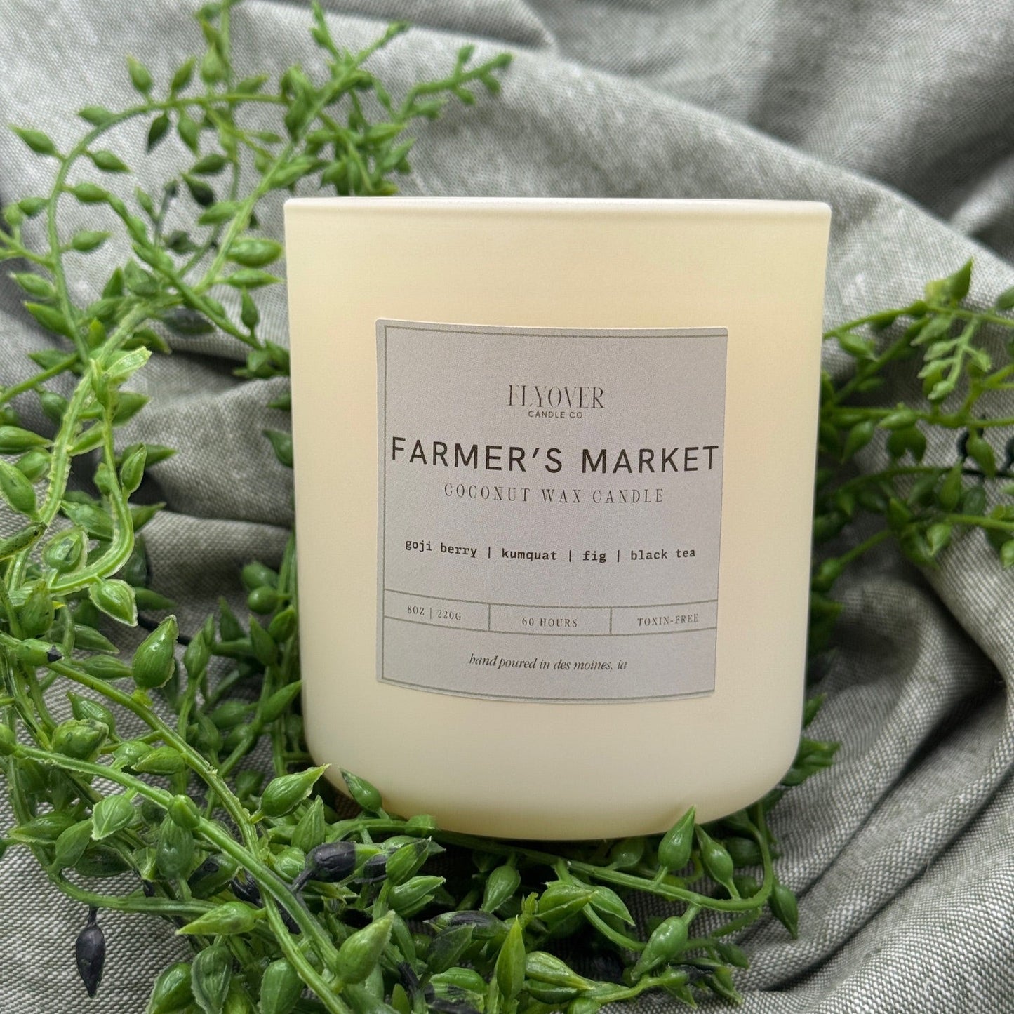 Candle labeled 'Farmer's Market' on a gray fabric background with green leaves.