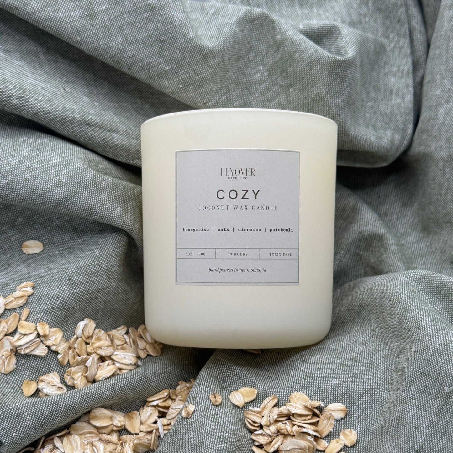 Candle labeled 'Cozy' on a gray fabric background with oats around