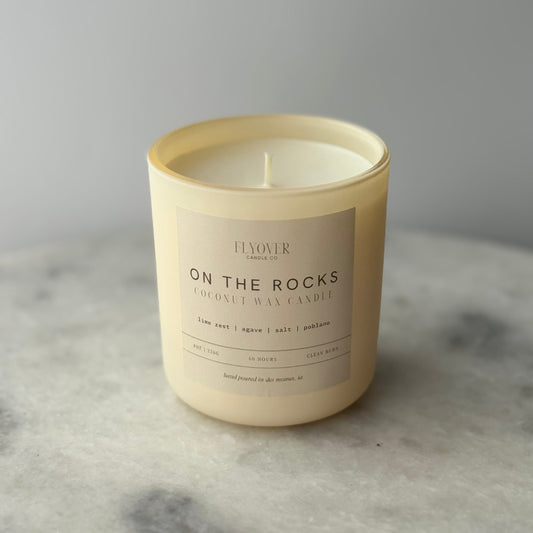 Candle labeled 'On the Rocks' by on a marble surface with a gray background