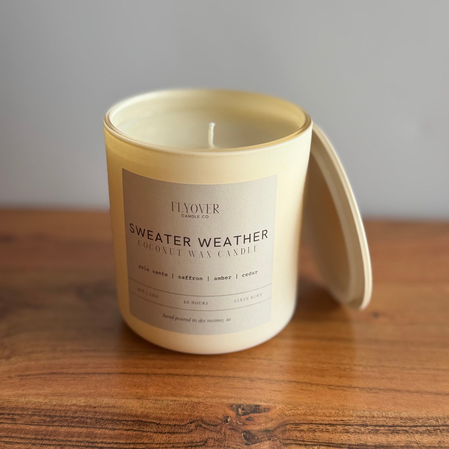 A cream-colored coconut wax candle in a matching ceramic jar with its lid resting beside it on a wooden table. The label reads 'FLYOVER Candles SWEATER WEATHER'.