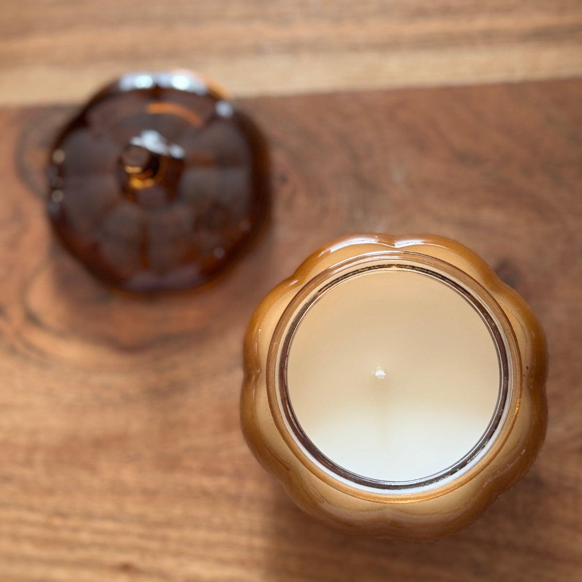 A close-up of a candle in a scalloped amber glass jar with its lid placed beside it on a wooden surface.