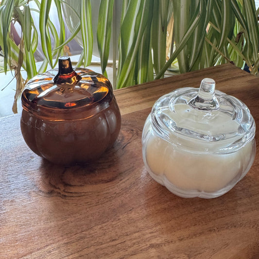 Two pumpkin-shaped glass containers with lids sit on a wooden tabletop, with green houseplants in the background. One container is amber brown and the other is clear glass containing a white candle or wax.