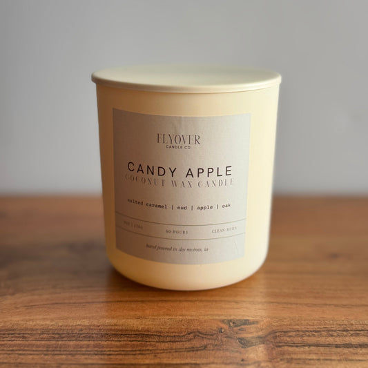 Ivory candle vessel with lid and label stating Candy Apple fragrance. Candle is placed on a wood table with white back drop. 