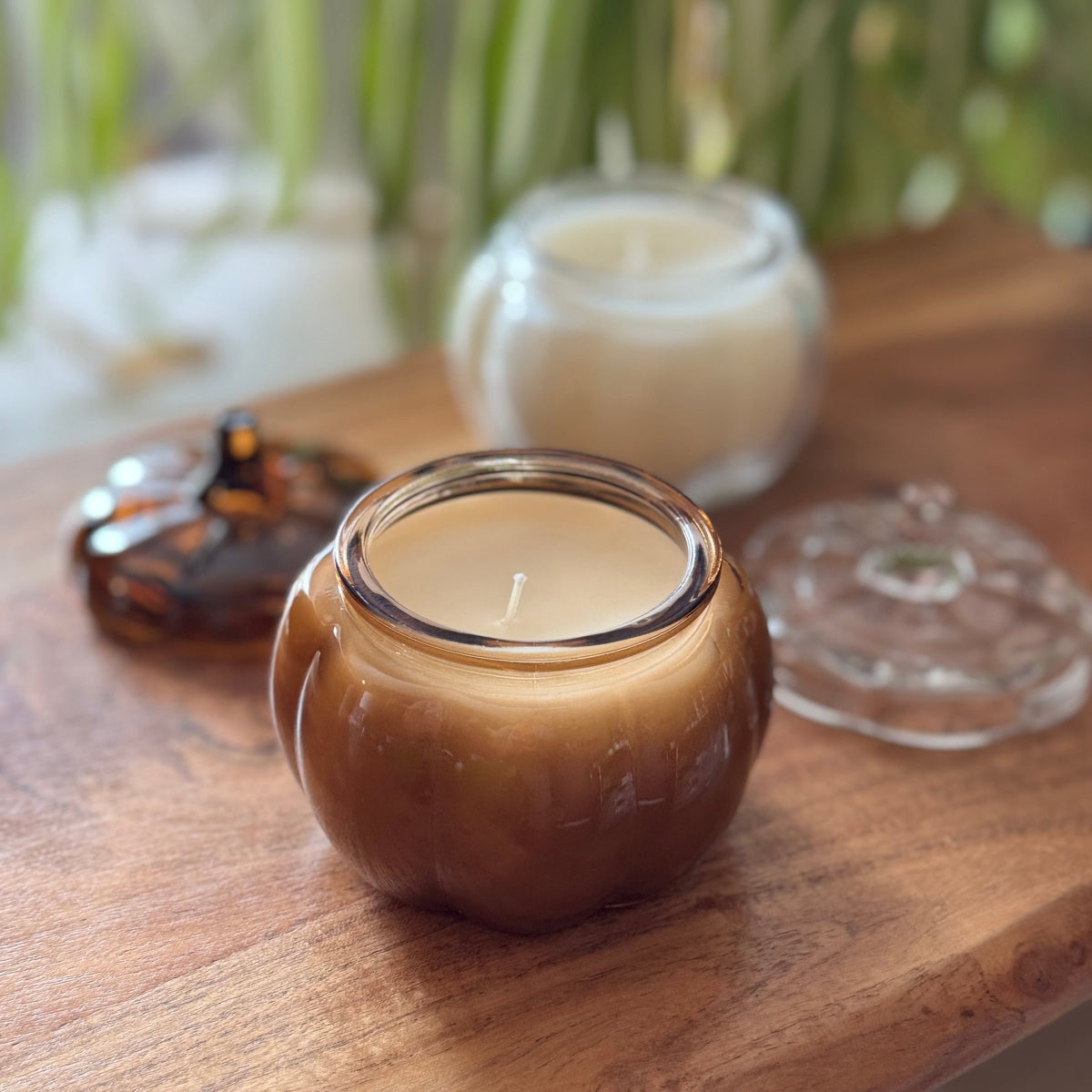 A pumpkin-shaped amber glass candle jar filled with cream-colored wax, shown on a wooden table with its clear glass lid and a small amber decorative piece in the background.