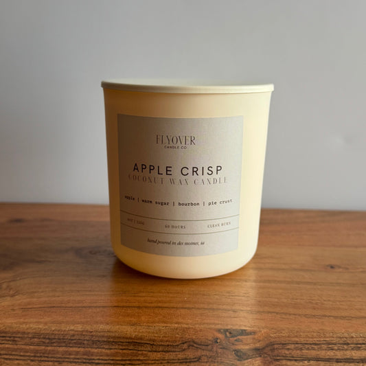 A large cream-colored coconut wax candle by Flower Candle in a white glass jar with a beige label reading 'APPLE CRISP'. The candle is placed on a rustic wooden table against a neutral gray wall.