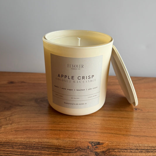 A large cream-colored wax candle in a matching open jar with its lid leaning against the right side, placed on a wooden tabletop against a plain gray wall. The front label reads 'FLYOVER' and 'APPLE CRISP COCOA-OIL WAX CANDLE'.