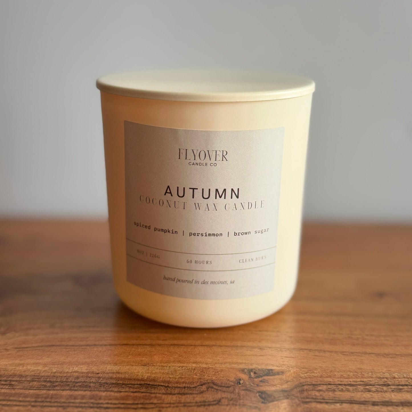 An ivory candle with the fragrance label Autumn and Flyover Candle Co. Candle is set on a wooden table with a white back drop. 