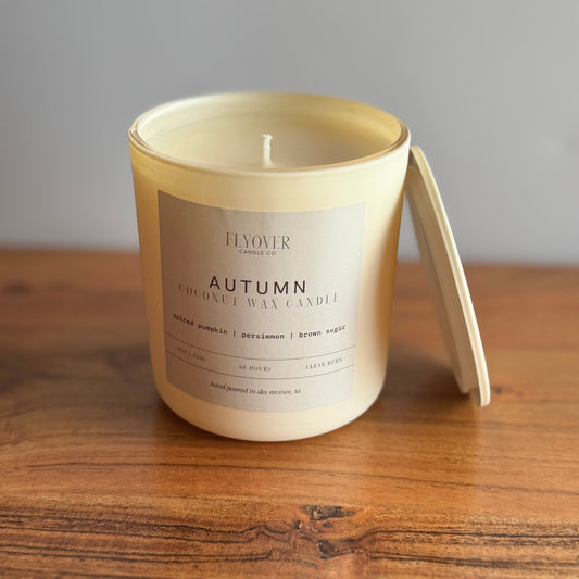 A creamy off white coconut wax candle in an ivory vessel with the fragrance label Autumn and Flyover Candle Co. Candle is placed on a wooden table with white back drop. 