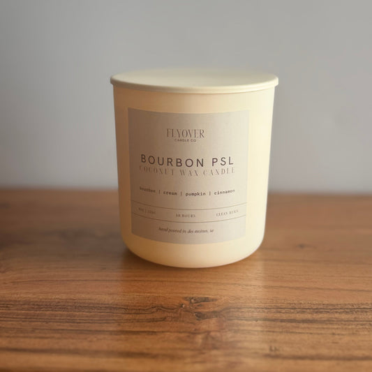 A large cream-colored coconut wax candle by Flower Candle in a white glass jar with a beige label reading 'BOURBON PSL'. The candle is placed on a rustic wooden table against a neutral gray wall.