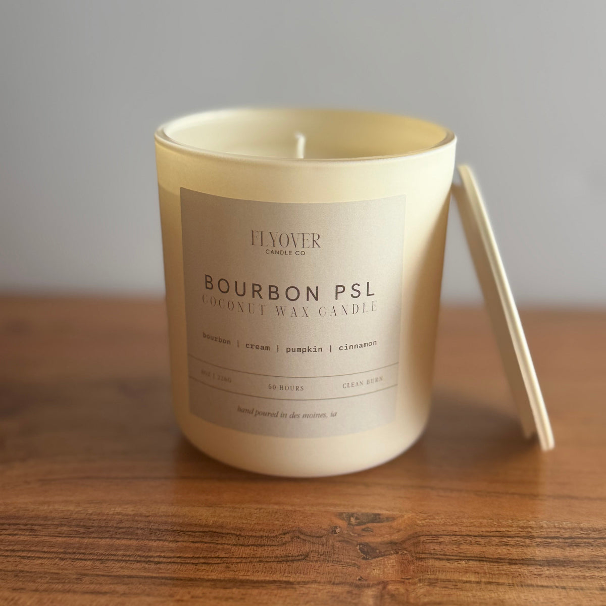 A cream-colored wax candle in a glass jar sits on a wooden table with its lid leaning against the right side. The label reads 'FLOVER CANDLE CO BOURBON PSL'.