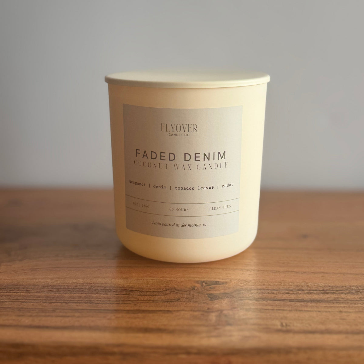 A cream-colored coconut wax candle in a matte white jar with a beige label reading 'FADED DENIM' by Flyover Candle Co, sitting on a wooden surface against a neutral gray background.