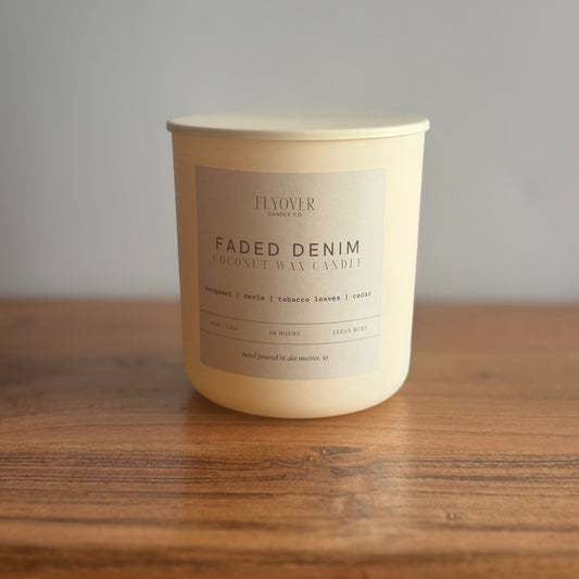 A cream-colored coconut wax candle in a matte white jar with a beige label reading 'FADED DENIM' by Flyover Candle Co, sitting on a wooden surface against a neutral gray background.