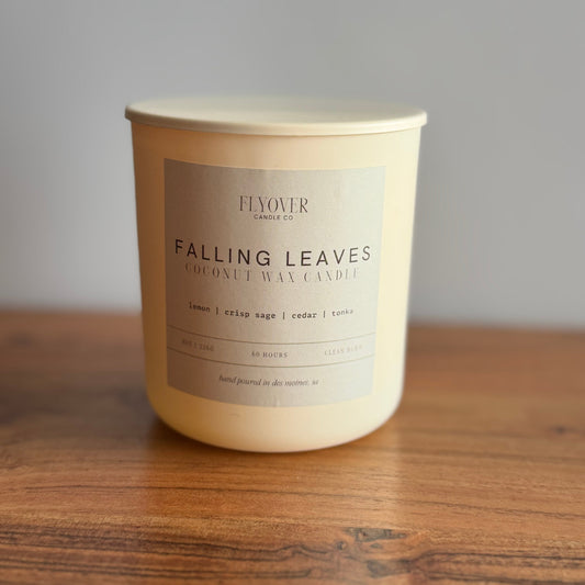 An ivory candle with the fragrance label Falling Leaves and Flyover Candle Co placed on a wooden table with white back drop. 