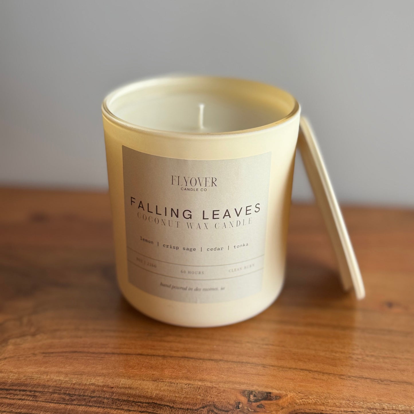 A cream-colored coconut wax candle in a glass jar with a matching lid leaning against the side, placed on a wooden table against a neutral gray background. The label reads 'FJOUR' and 'FALLING LEAVES'.