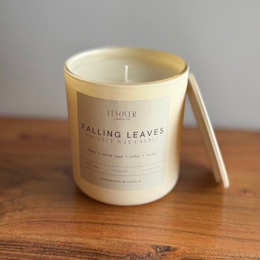 A cream-colored coconut wax candle in a glass jar with a matching lid leaning against the side, placed on a wooden table against a neutral gray background. The label reads 'FJOUR' and 'FALLING LEAVES'.