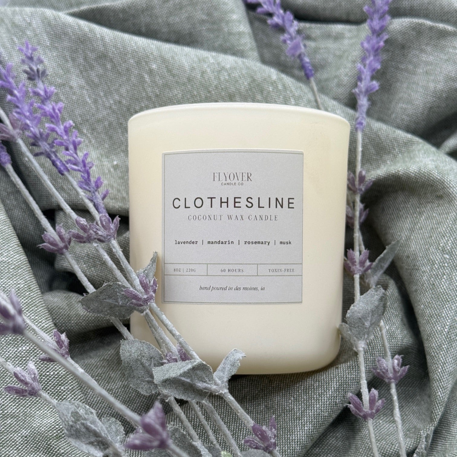 Candle labeled 'Clothesline' surrounded by lavender on a textured fabric background