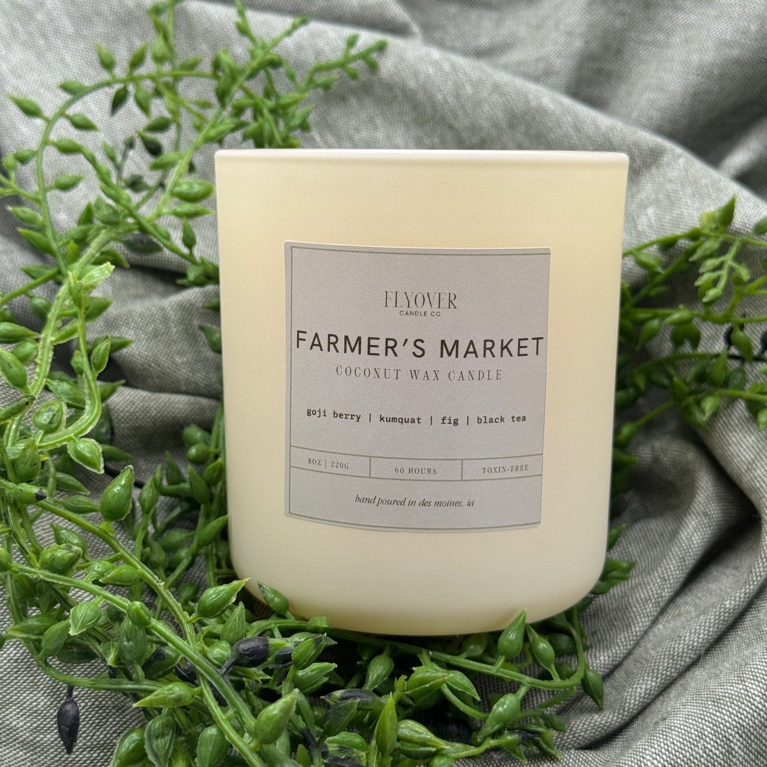 Candle labeled 'Farmer's Market' on a gray fabric background with green leaves.