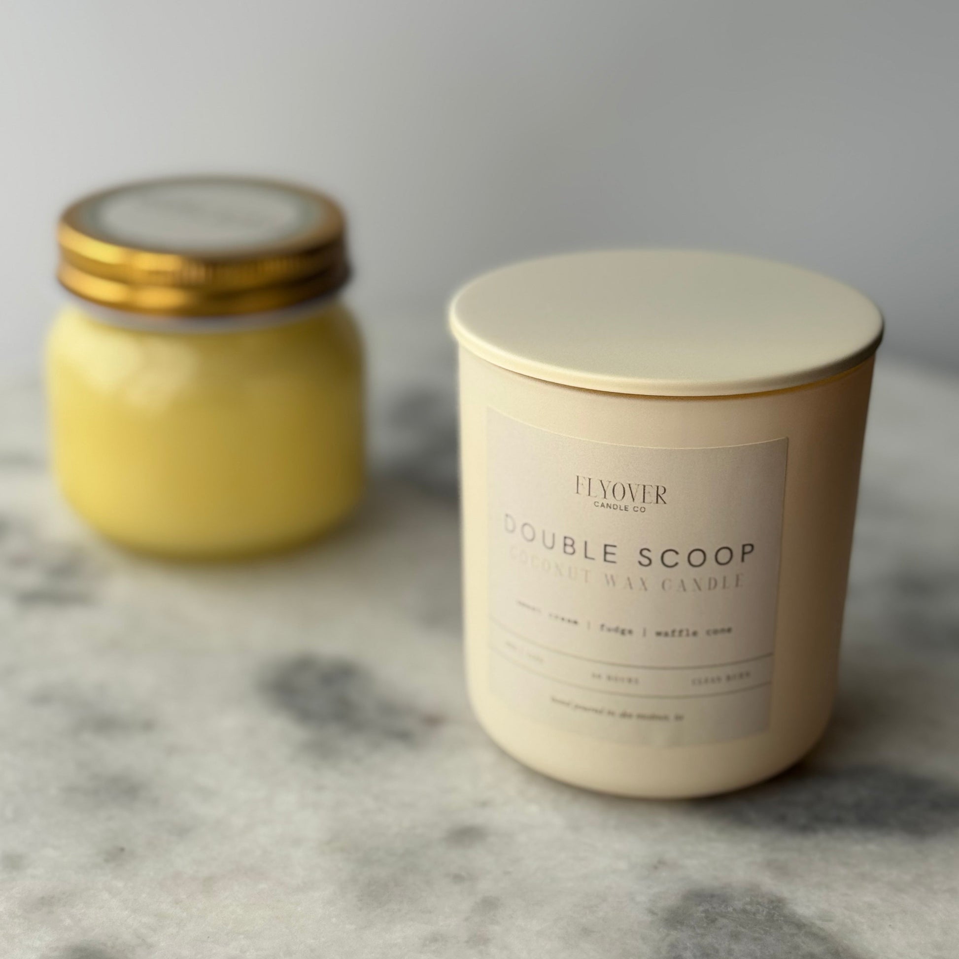 Two candles, one small and yellow, the other large and white with a label, on a marble surface.