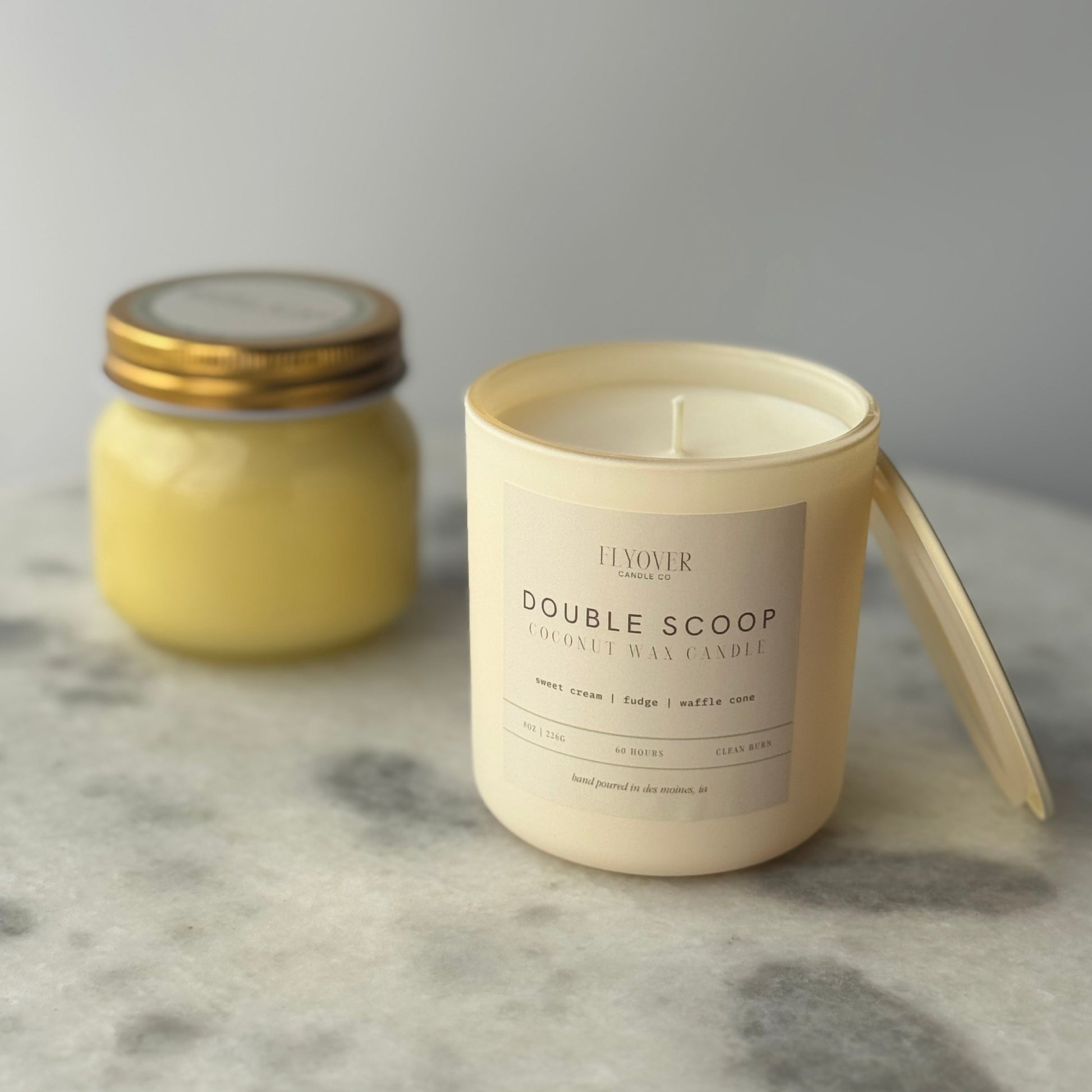 Two candles, one in a jar and one in a white container with a label, on a gray surface.