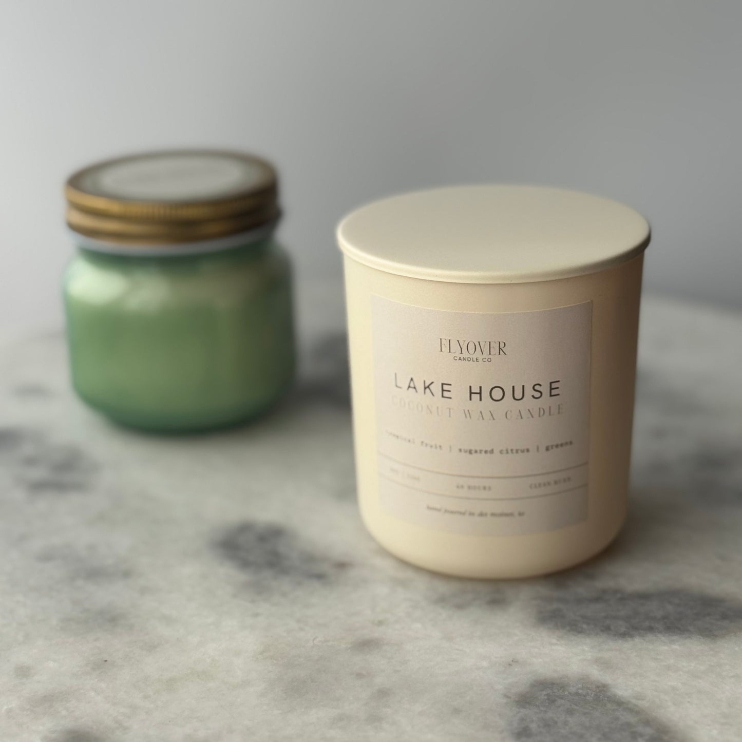 Two candles, one green and one beige with 'Lake House' text, on a marble surface.