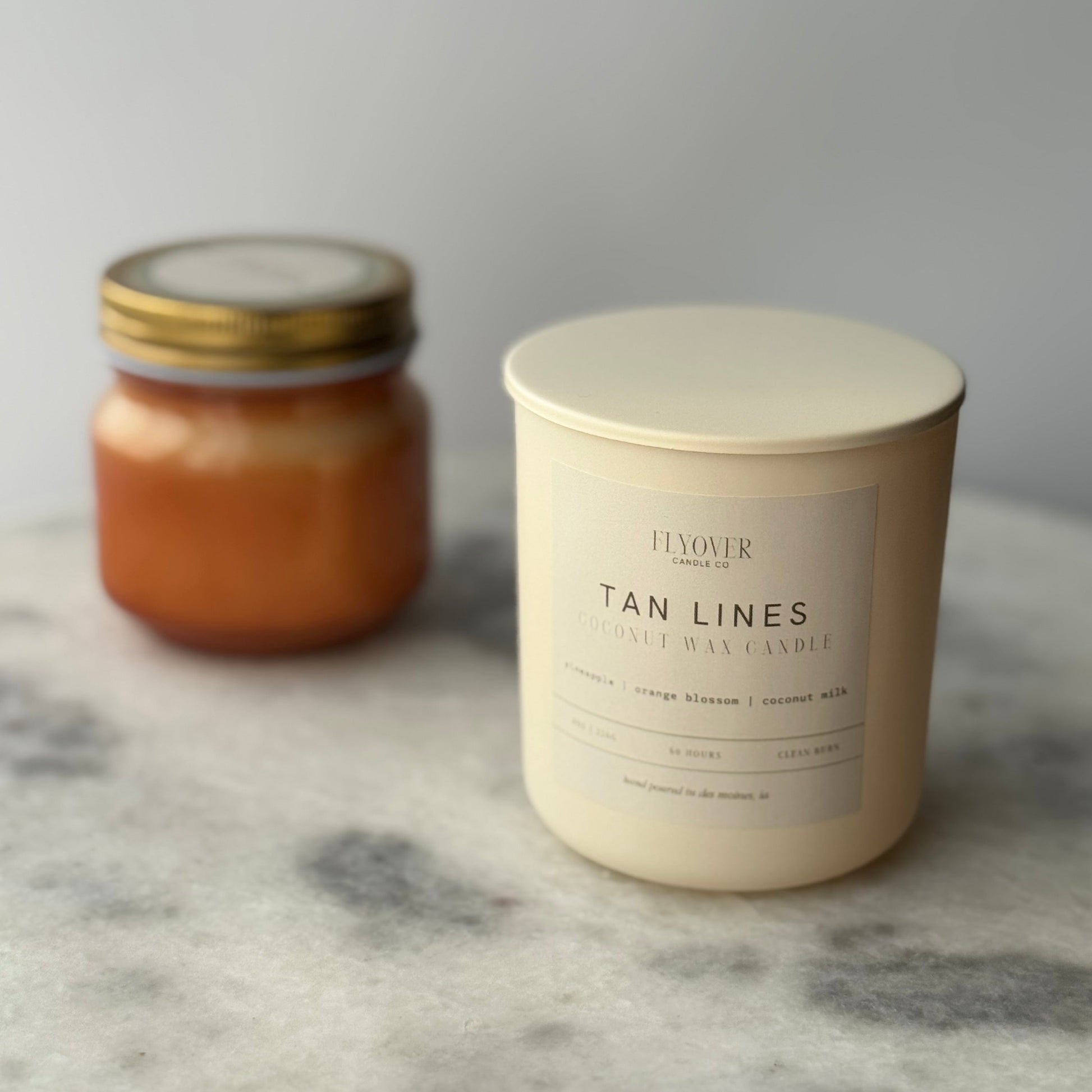 Two candles, one in a glass jar and one in a white container with a label, on a marble surface.