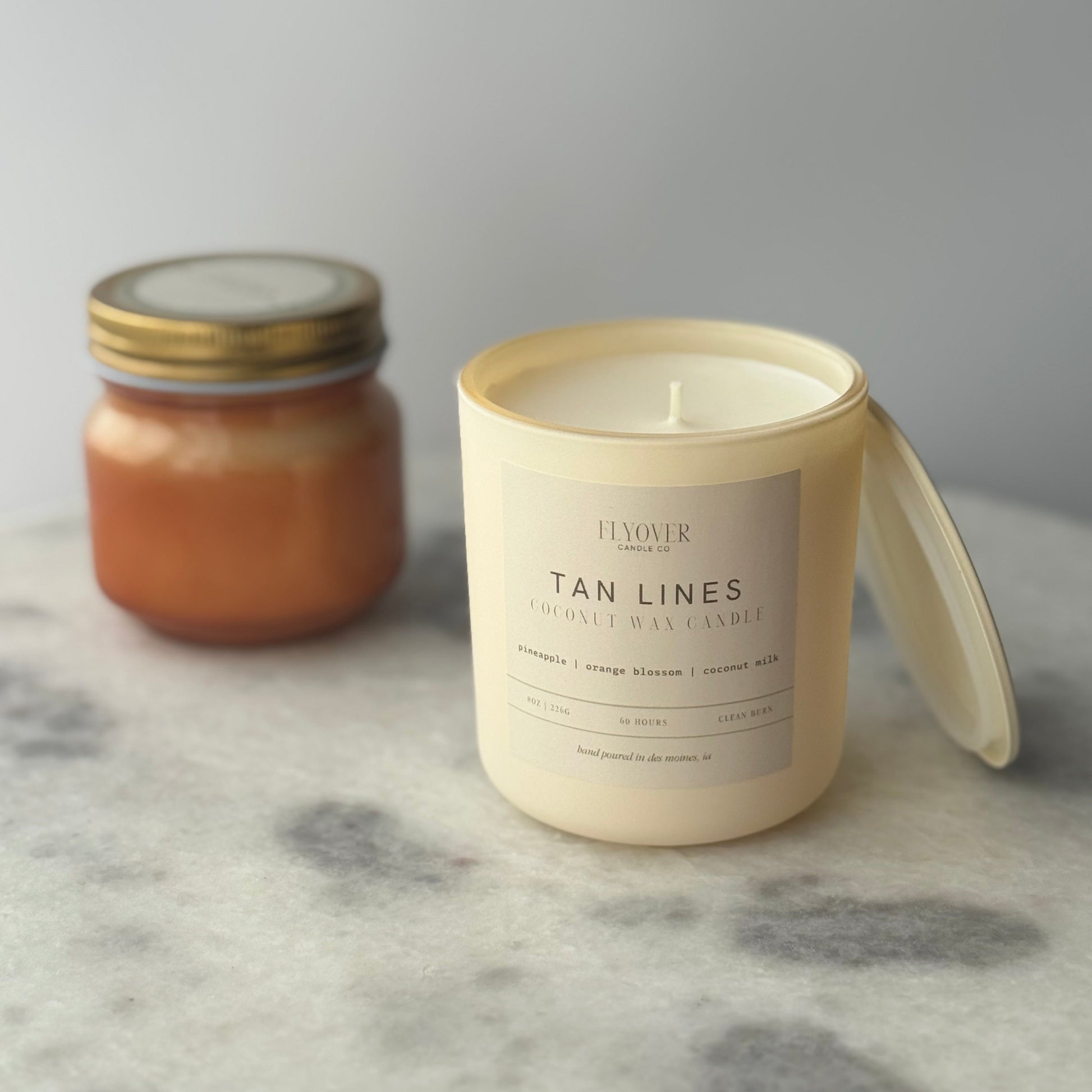 Two candles on a marble surface, one in a jar and one in a white container with a label.