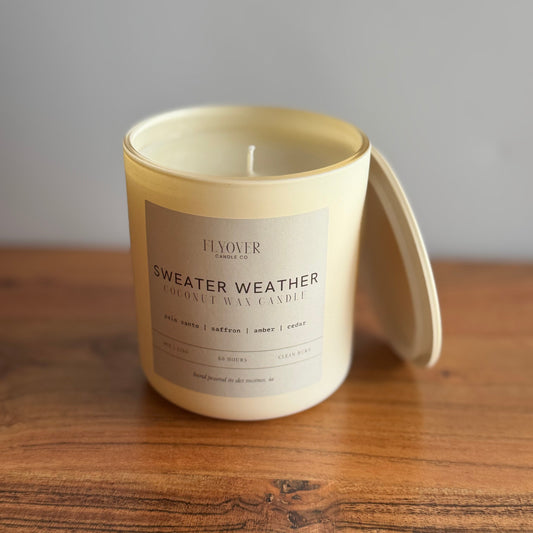 A cream-colored coconut wax candle in a matching ceramic jar with its lid resting beside it on a wooden table. The label reads 'FLYOVER Candles SWEATER WEATHER'.