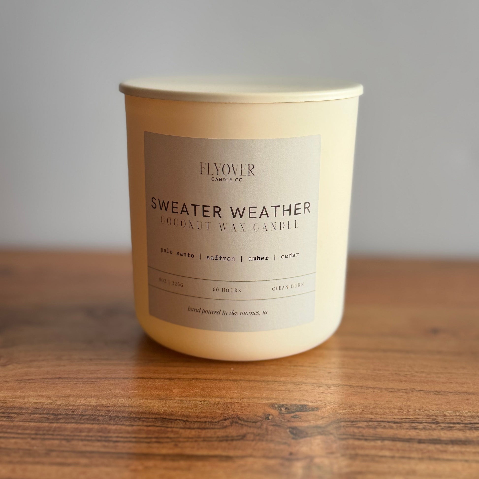 An ivory coconut wax candle with the fragrance label Sweater Weather and Flyover Candle Co placed on a wood table with white back drop. 