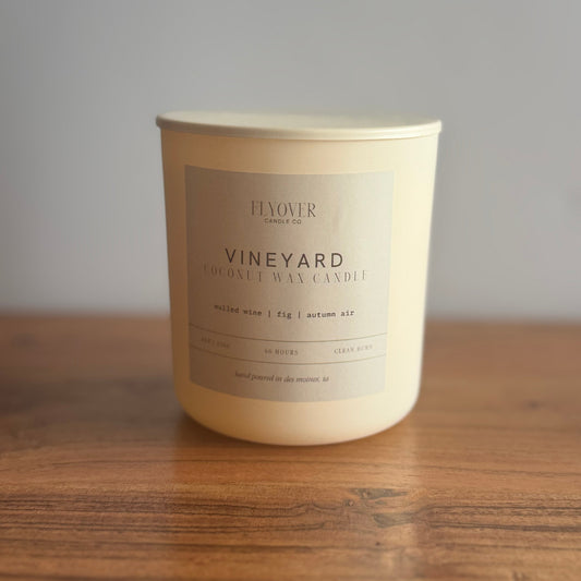 An ivory candle with the fragrance label Vineyard and Flyover Candle Co placed on a wood table with white back drop. 