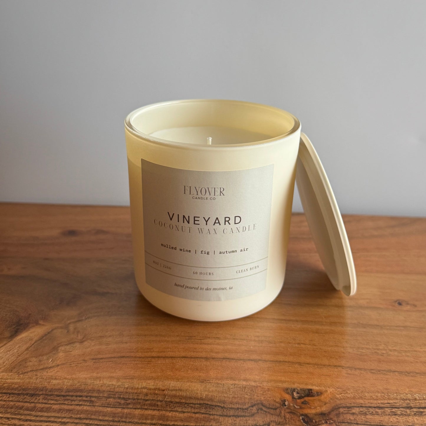 A cream-colored jar candle with its lid leaning against it, placed on a wooden tabletop against a plain gray wall. The front label reads 'FLOVER' and 'VINEYARD COCONUT WAX CANDLE'.